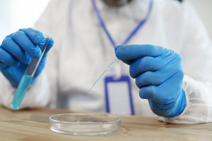 Wissenschaftler in Schutzkleidung führt ein Experiment durch, indem er eine Flüssigkeit aus einem Reagenzglas mit einer Pipette in eine Petrischale tropft.